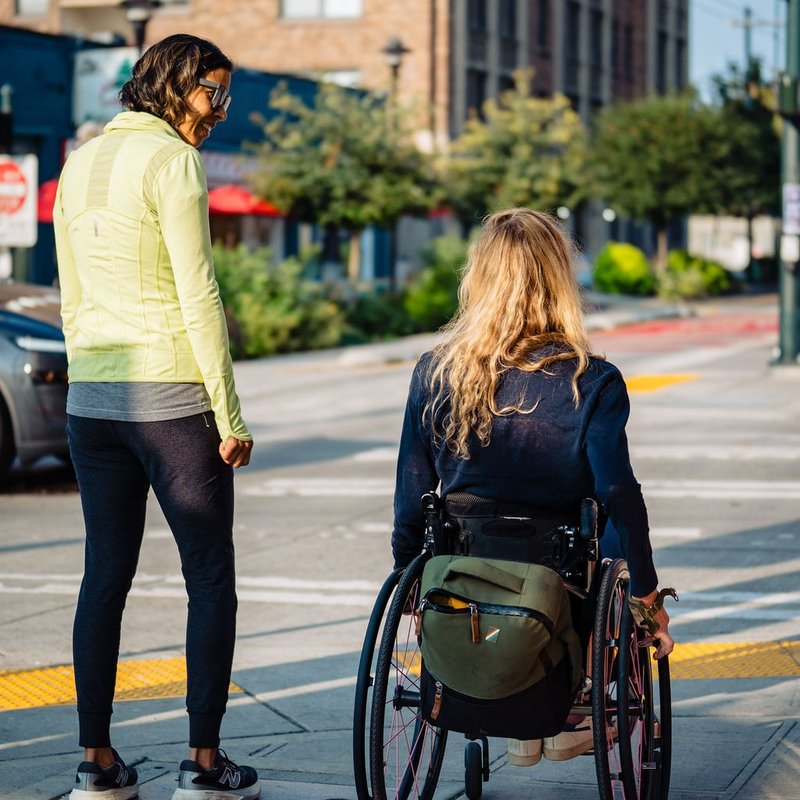 Two women waiting to cross the street, one standing and smiling. The other in a wheelchair with a backpack, face not visible.