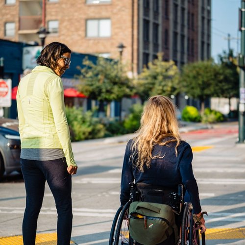 Two women waiting to cross the street, one standing and smiling. The other in a wheelchair with a backpack, face not visible. A local restaurant with table umbrellas indicating outdoor seating is across the intersection.