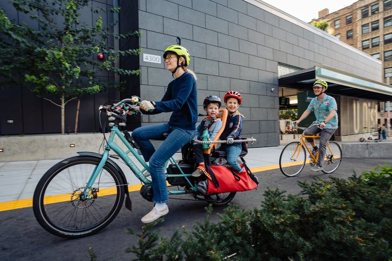 People biking while wearing helmets in a protected bike lane in front of U District Station. Mom is riding a cargo bike with two smiling kids on the back. Dad follows behind.