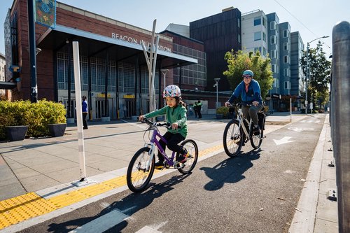 Child and adult biking together in a bike lane next to the sidewalk in front of the Beacon Hill light rail station. On the other side of the bike lane, a railing separates it from a bus island (not fully visible) where people wait for the bus.”