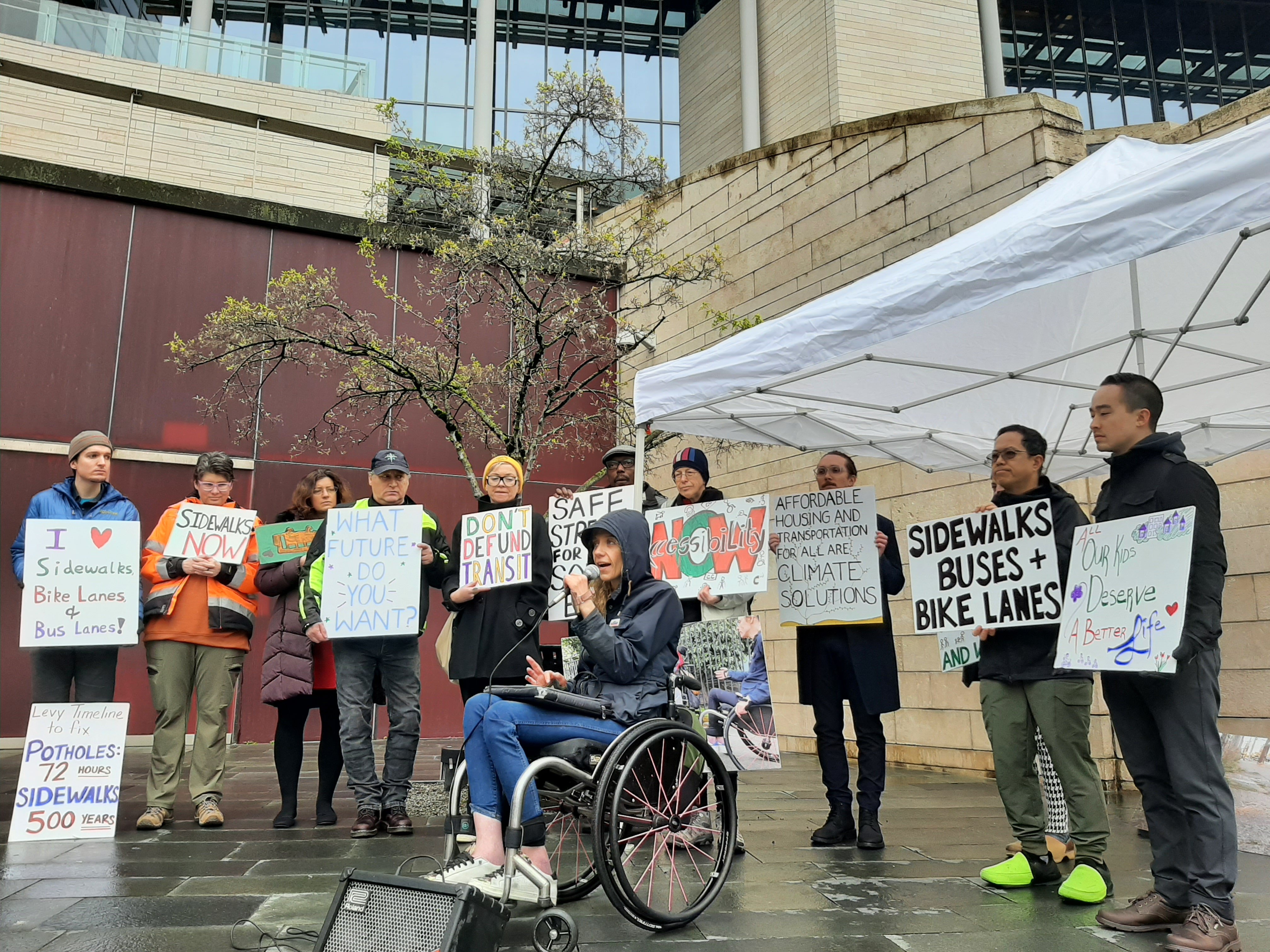 A woman sitting in a wheelchair speaks into a microphone in front of a row of people wearing raincoats and holding signs promoting and demanding sidewalks, buses, and bike lanes.