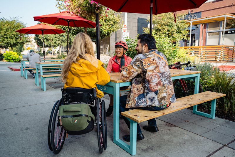 People sitting and talking at a picnic table on a sidewalk. Two are seated on the bench, one is using a wheelchair.