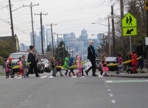 A row of small children cross a street in front of a "pedestrian crossing" sign.