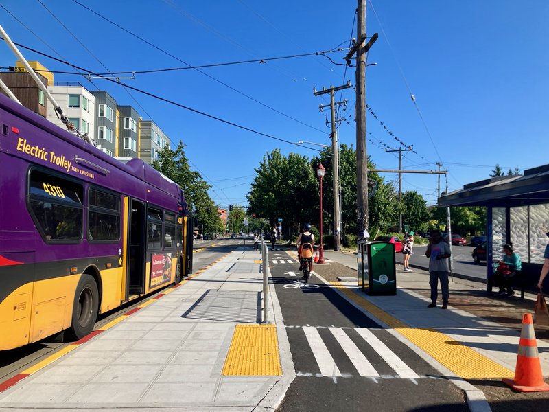 Trolly bus on Beacon Hill with doors opening to a floating island. A bicylist passes by in the bike lane while people wait at the bus shelter for the next bus