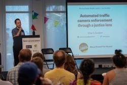 Ethan Campbell speaking at a lectern in front of an audience. Slide on large screen says: Automated traffic camera enforcement through a justice lens.