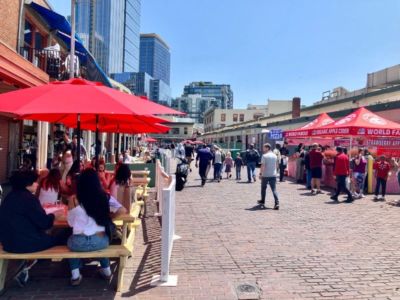 Pike Place with people walking in the street while it is closed to regular vehicle traffic. People are seated at picnic tables under umbrellas at the end of the street on the left. On the right, farmers market booths are busy with shoppers.