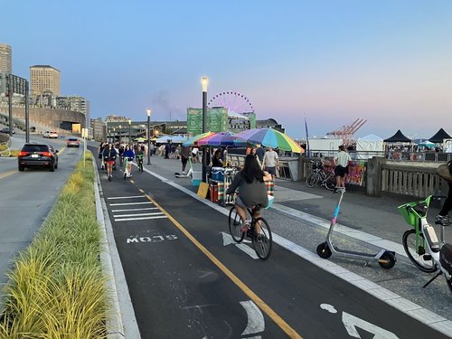 People biking and riding scooters in the new protected bike lane along Alaskan Way on the downtown waterfront. Sidewalk drink vendor with colorful umbrellas along the sidewalk. Downtown, waterfront, port skyline visible in the distance.