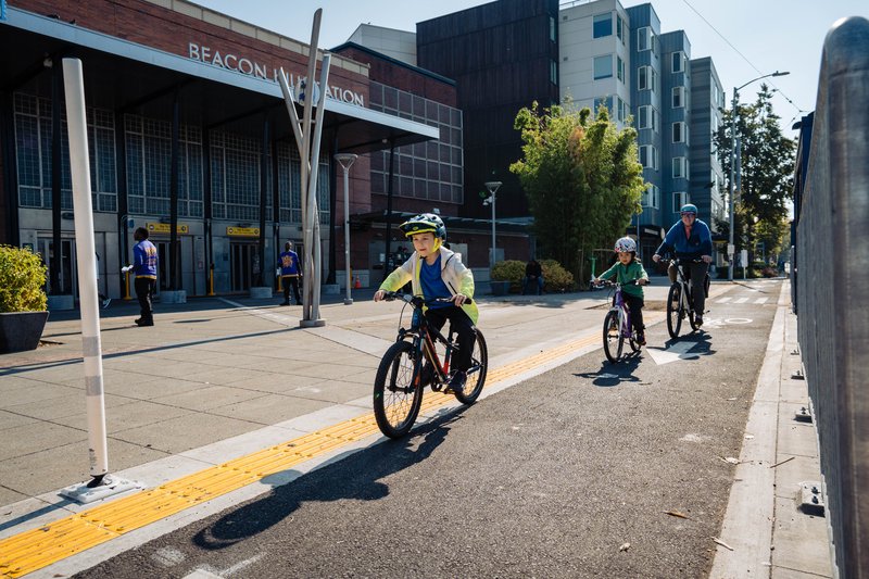 Two kids biking with an adult biking behind them in a protected bike lane between the sidewalk in front of Beacon Hill Station and a bus stop, not fully visible.