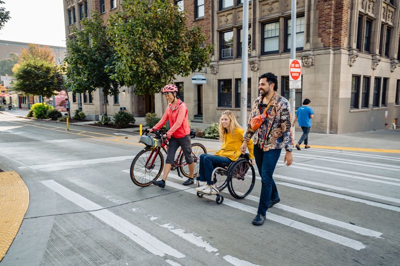 Three people crossing a crosswalk in the U District together, smiling: one walking a bike, one using a wheelchair, and one walking.
